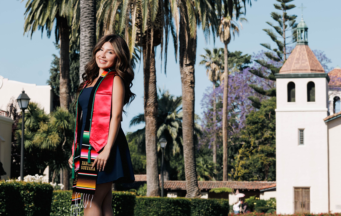 A young woman in her graduation stoles stands in front of the Mission Church.
